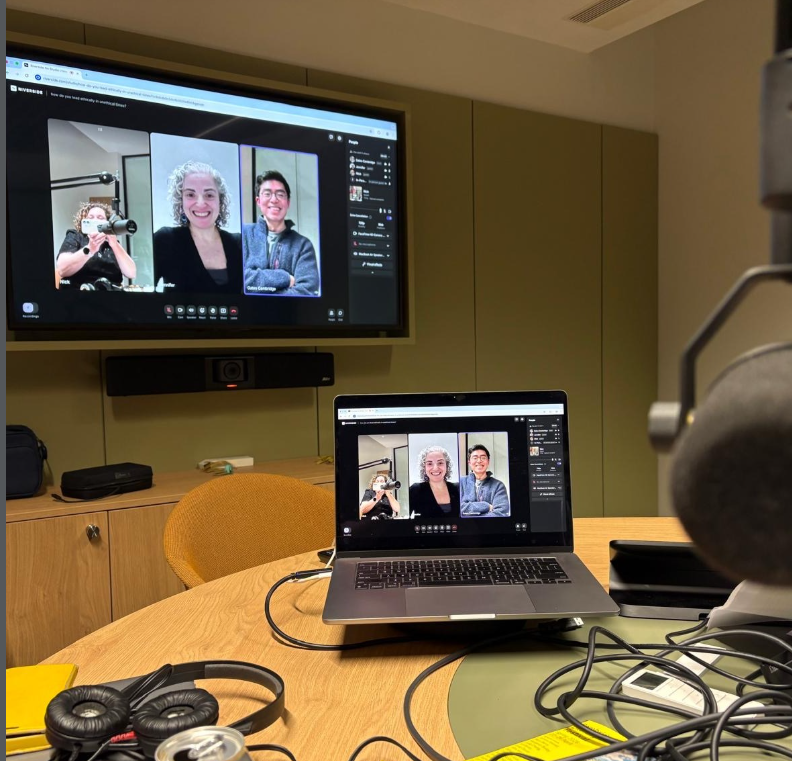Microphone in foreground and laptop on desk with three people on it and screen at the back on a wall with three people on it