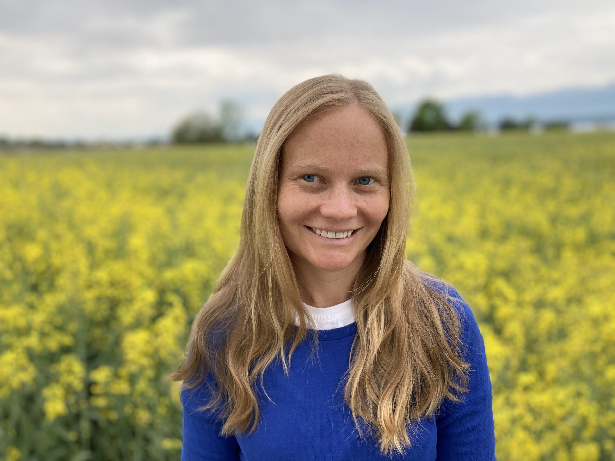 Woman with long hair in blue top in front of field of yellow flowers